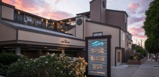 Exterior view of the Wave Street Inn in Monterey, California, with a sign featuring illuminated blue wave graphics and text "Wave Street Inn, Inns of Monterey", taken at dusk under a colorful sky, with sidewalk and lush landscaping in the foreground.