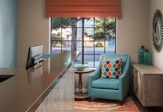 Hotel reception area featuring a brown countertop, a teal armchair with patterned pillow, round wooden side table, large window overlooking a scenic view, and decorative mirror and bottles on a wooden cabinet.