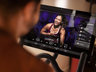 Person using a stationary exercise bike, watching a woman instructor on the screen, who is smiling and flexing her arm; screen displays workout metrics and a leaderboard.