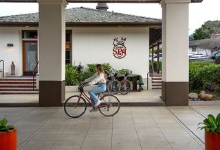 A young woman rides a red vintage bicycle on a tiled walkway in front of a building with a "Casa Spa" sign; other bicycles are parked nearby.