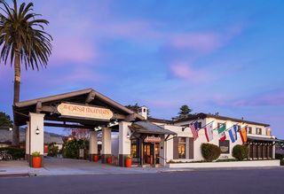 Exterior view of Casa Munras Garden Hotel & Spa at dusk, featuring a porte-cochere with a sign, the main building with the Esteban Restaurant sign and flags of several countries, and a palm tree in the foreground.