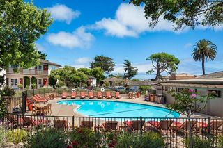 Exterior view of a hotel swimming pool with blue water, surrounded by lounge chairs, tables, and a black metal fence, with a two-story building featuring red doors and windows in the background, all under a bright blue sky with scattered clouds.