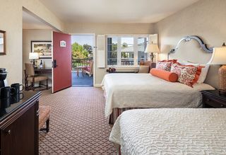 Hotel room featuring two queen beds, an open door leading to a balcony, a desk with chair and coffee station, with neutral walls, patterned carpet, and decorative pillows on the beds.