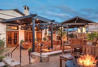 Outdoor restaurant patio with wooden tables and chairs under a dark pergola with string lights, heaters, and a fireplace at dusk, with an adjacent building and blue sky.