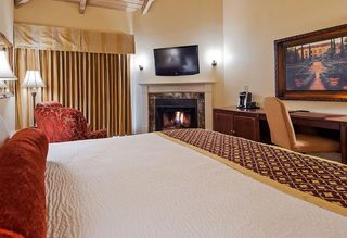Hotel room interior featuring a bed with decorative bedding, a lit fireplace beneath a wall-mounted TV, a sitting area with a patterned armchair, a desk with a painting above it, and golden curtains.