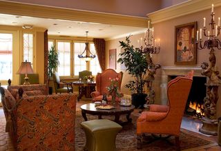 Cozy, traditionally styled living room with a fireplace, ornate furniture, and warm lighting. An upholstered sofa and two patterned wingback chairs surround a round marble-topped coffee table with wine and snacks. A lit fireplace is flanked by candle-lit sconces and statues, with artwork above the mantel. In the background, a dining area features a round table, chairs, and a chandelier. The room is softly lit with warm tones.
