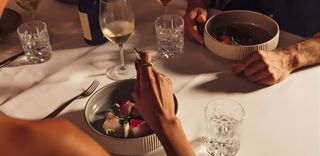 Close-up of a dinner table setting with people, white tablecloth, and various dishes. A person's hand reaches for a dish of food, illuminated by soft light. Other glasses of wine, water, and another person can be seen at the table.