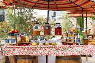 A Bloody Mary bar set up outdoors under striped umbrellas, featuring three large glass beverage dispensers of carrot, classic bloody, and clamato juice, surrounded by jars of pickles, olives, pickled eggs, and various condiments.