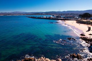 Coastal view of a calm blue ocean meeting a sandy beach and a long pier with boats. Mountains and clear blue sky are in the background.