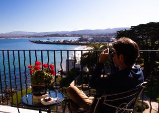 A man using binoculars views a coastal scene from a balcony, with a view of boats in the harbor.