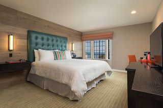 A hotel bedroom with a large bed featuring a tufted turquoise headboard, white bedding, and a striped decorative pillow, situated against a wood-paneled wall with sconce lighting, a window with a view of the ocean, a desk, and a television.