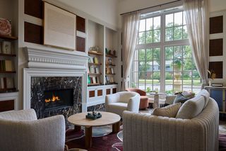 Royal Park Hotel Library, featuring a lit marble fireplace with a lit fire surrounded by built-in bookshelves and artwork. The space is furnished with two armchairs, a curved sofa, and a round wooden coffee table.