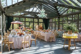 Elegant event setup inside a sunlit conservatory, featuring multiple tables adorned with floral centerpieces, set for dining, and surrounded by wooden chairs, with an accent table showcasing refreshments.