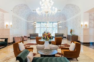Elegant hotel lobby with a large chandelier, ornate floral arrangements, seating areas with velvet chairs, and a marble floor with a patterned rug.