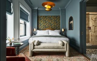 Luxurious hotel bedroom with a large bed against a floral wallpaper headboard, a bench at the foot of the bed, a vintage rug, and an en-suite bathroom visible through a doorway on the right.