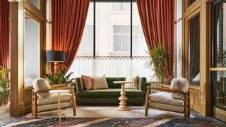 Interior of a warm lobby featuring a green velvet sofa with decorative pillows, two matching armchairs, wooden side table, large window framed by rust-colored velvet curtains, and a floor lamp.