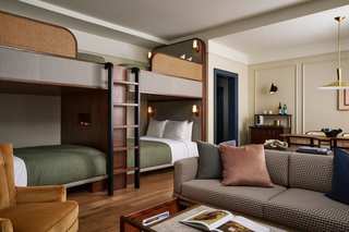 Interior shot of a hotel room featuring two custom bunk beds. The room also includes a sofa with throw pillows, a small table, an armchair, a dining area, and a bar area. The room has cream-colored walls, gold lighting, and dark wood accents.
