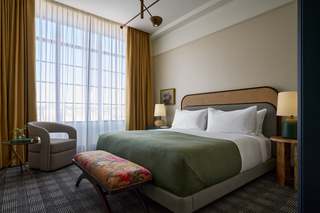 Hotel bedroom with a large bed covered with white bedding and a green throw, and a floral bench at the foot of the bed. A patterned armchair and small side table with a lamp sit to the left of the bed, near a large window with tan and sheer curtains.