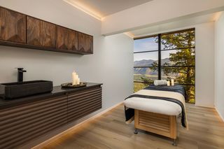 Spa treatment room with a massage table, a stone sink, and a large window overlooking a mountainous forest landscape.