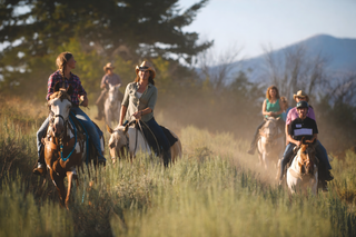 A group of people on horseback rides along a dusty trail through a dry, grassy landscape with trees and hills in the background.
