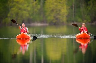 Two individuals in orange kayaks paddle across a calm lake, with lush green trees reflected in the water.