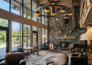 A rustic lodge interior with a large stone fireplace, mounted animal heads, a grand piano, and comfortable seating arranged around a rug, all under a large, ornate chandelier.