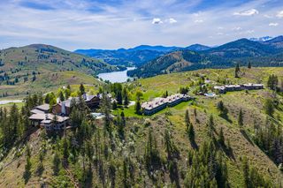 Aerial view of a Sun Mountain Lodge nestled on a hillside overlooking a serene lake and rolling, tree-covered mountains under a bright, partly cloudy sky.