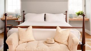 A four-poster bed with white linens is flanked by two small wooden desks, each with a lamp and vase of flowers. In front of the bed is a pale yellow tufted bench with two decorative pillows and a straw hat.