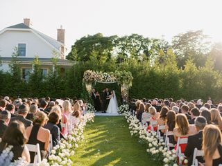 An outdoor wedding ceremony takes place in a grassy yard in front of a white house, with guests seated on white chairs lining a path decorated with white roses, and a floral archway centered behind the wedding party.