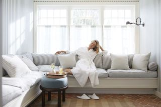 A blonde woman wearing a white robe relaxes on a striped L-shaped couch in a room with blue and white striped wallpaper and a large window with sheer curtains.