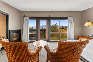 Two large wicker chairs face a round wooden coffee table in a cozy room with a lit fireplace, white curtains, and a large window overlooking a scenic landscape with mountains and trees.