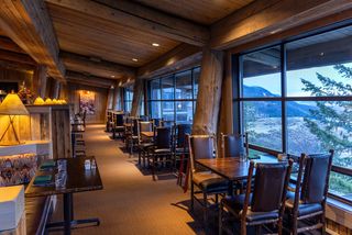 Rustic restaurant dining room with log beams, wooden tables, and leather chairs, overlooking a scenic mountain landscape at dusk.
