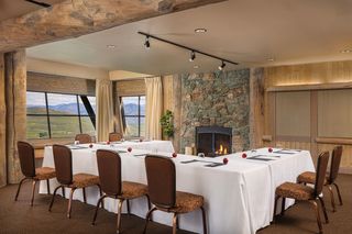 A U-shaped meeting table covered in white tablecloths, set with glasses, papers, and red apples, surrounded by upholstered chairs in a room with a large stone fireplace, wooden beams, and windows overlooking a scenic mountain landscape.