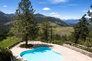 A swimming pool on a concrete patio overlooking a lush green valley and distant snow-capped mountains.
