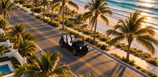 A black open-top vehicle drives down a coastal road lined with palm trees and a sandy beach, with the ocean waves in the background under warm sunlight.